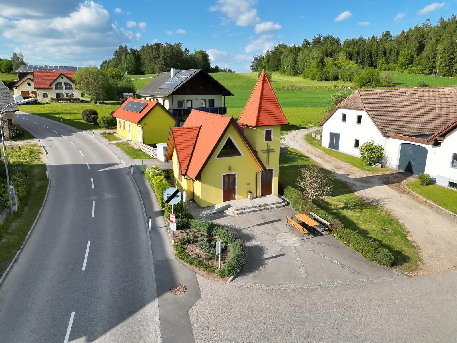 Luftaufnahme eines kleinen Rastplatzes mit gelber Kapelle und Sitzbank an einer Landstraße.