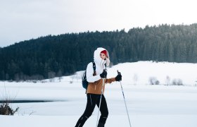 Eine Person beim Skilanglauf auf einem verschneiten Feld vor einem Wald.