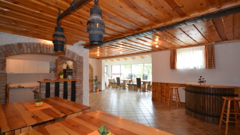 Innenansicht eines rustikalen Gastraums mit Holztischen und -decken, Barbereich und großen Fenstern.