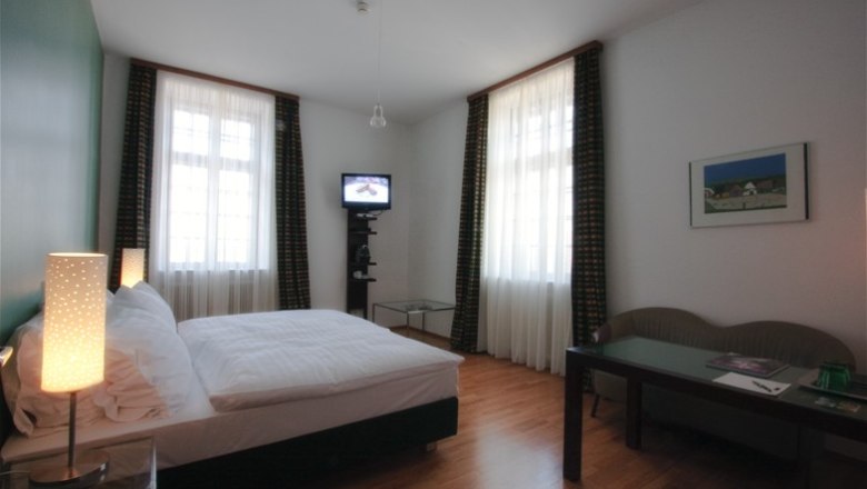 Ein Hotelzimmer mit Doppelbett, zwei Fenstern, einem kleinen Fernseher und einem Tisch mit Stuhl.