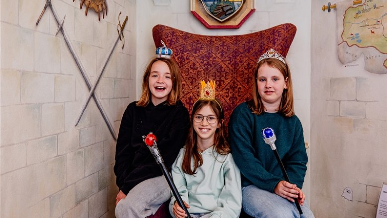 Drei Kinder mit Kronen und Zeptern sitzen auf einem Thron in einem m&auml;rchenhaften Raum.