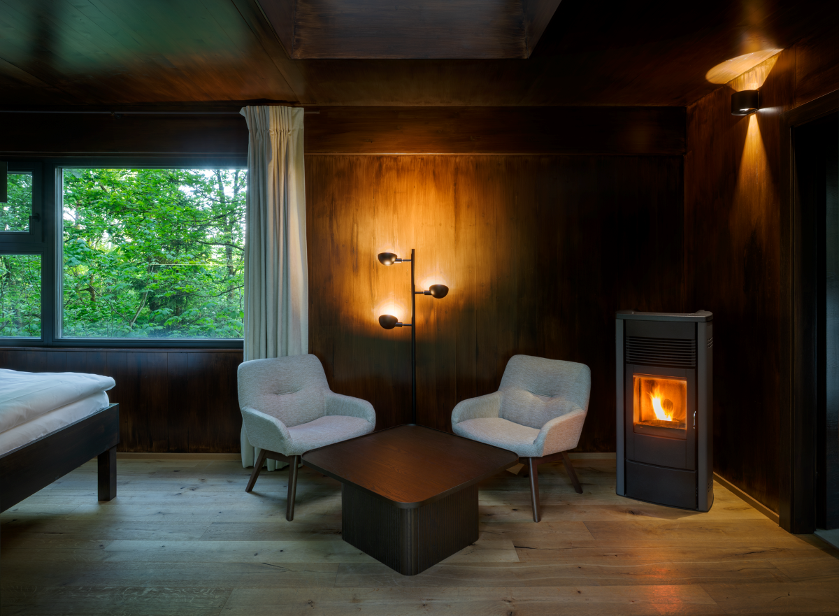 Innenraum eines Baumhauses mit zwei Sesseln, Tisch, Kamin und großem Fenster mit Blick ins Grüne.