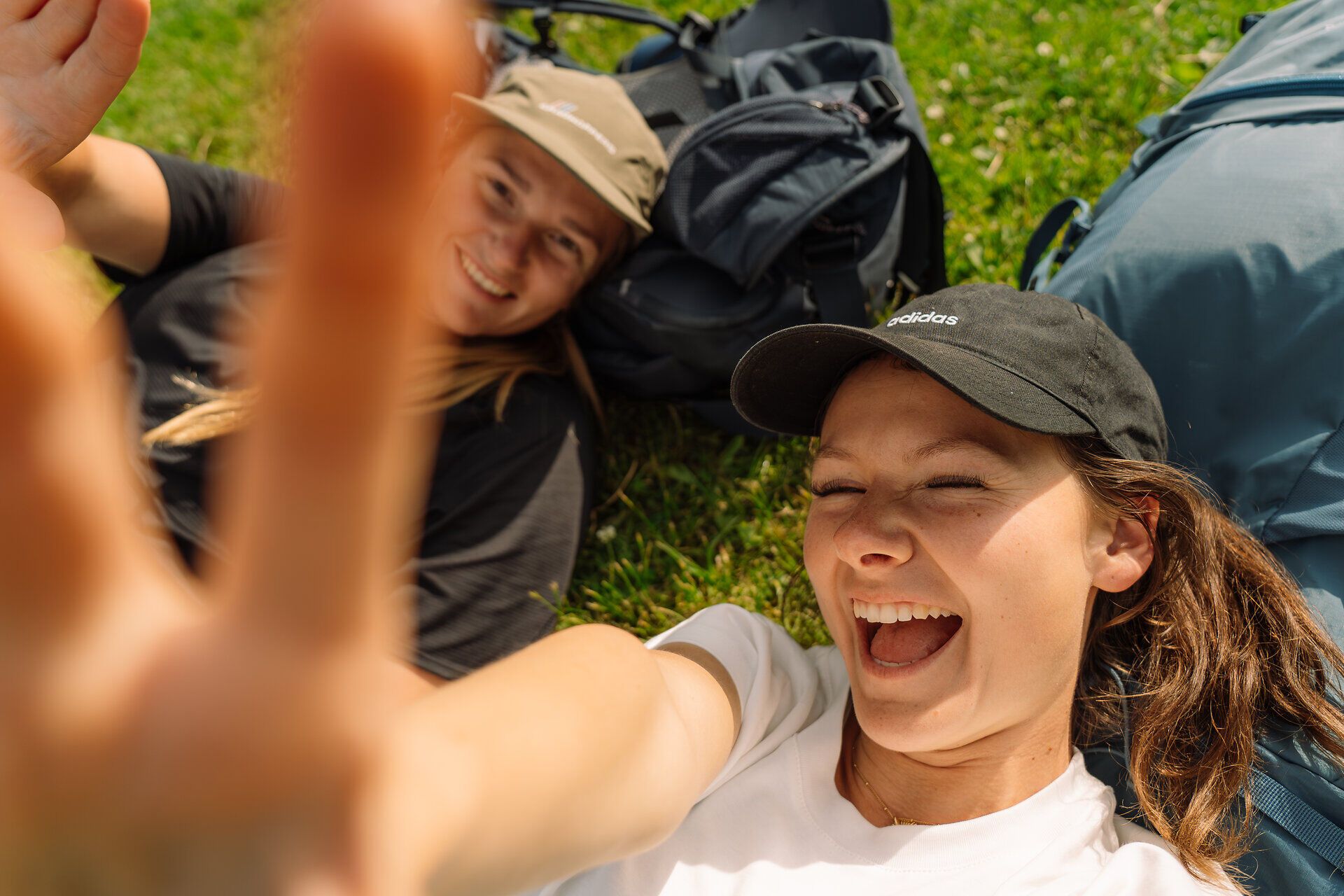 Zwei Frauen lachen in die Selfiekamera, während sie auf ihren Rucksäcken auf einer Wiese liegen
