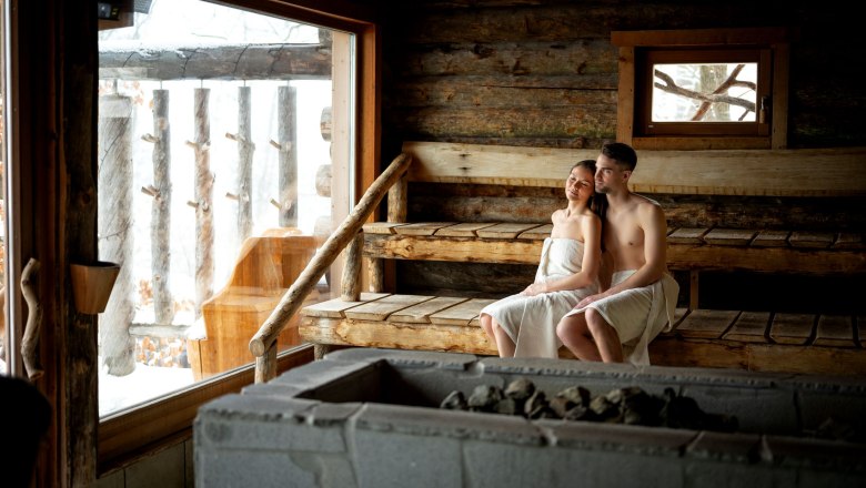 Ein Paar sitzt in einer Sauna aus Holz, eingewickelt in Handtücher, mit Blick nach draußen auf eine verschneite Landschaft.