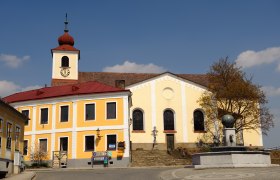 Kirche mit gelber Fassade und Turm mit rotem Dach auf einem Hauptplatz.