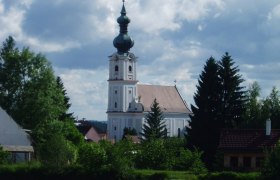 Kirche in Kirchberg am Walde mit Teich im Vordergrund und bewölktem Himmel.
