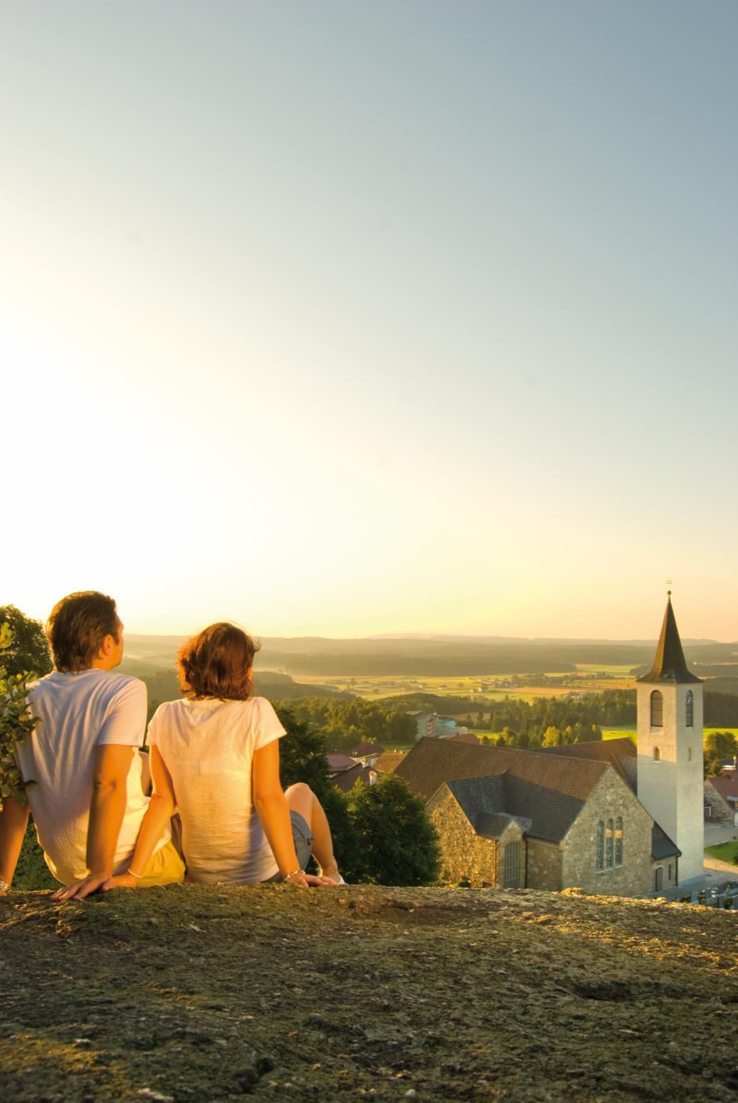 Zwei Personen sitzen auf einem Felsen und blicken auf eine Kirche und eine weite Landschaft bei Sonnenuntergang.