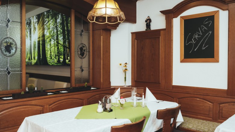 Gemütlicher Gastraum mit Holzmöbeln, Tisch mit grüner Tischdecke, Kerzen und Dekoration. Fenster mit Waldmotiv und Tafel mit Schriftzug 'Servas'.