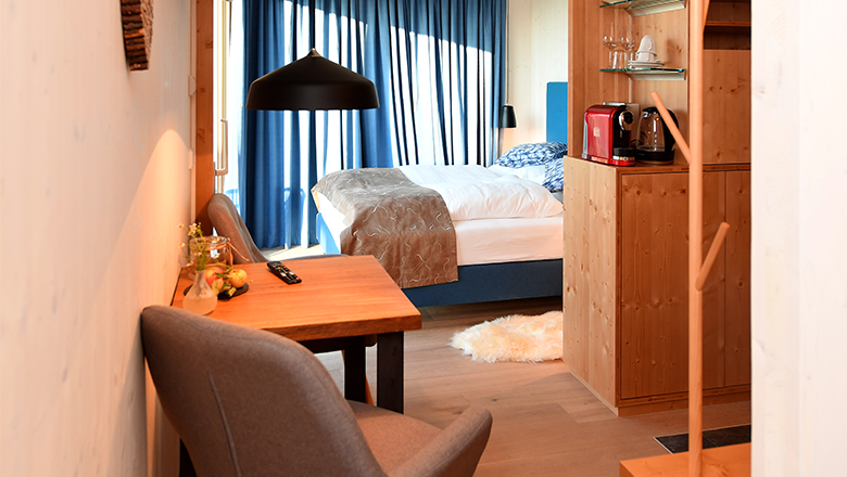 Gem&uuml;tliches Hotelzimmer mit Holzm&ouml;beln, einem Bett, Tisch und St&uuml;hlen. Eine Kaffeemaschine steht auf einem Regal. Blaue Vorh&auml;nge am Fenster.