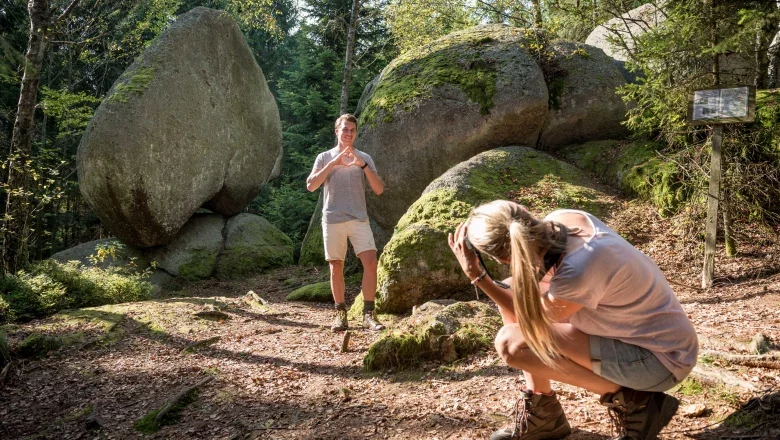 Ein Mann posiert vor einem gro&szlig;en herzf&ouml;rmigen Felsen im Wald, w&auml;hrend eine Frau ein Foto von ihm macht.