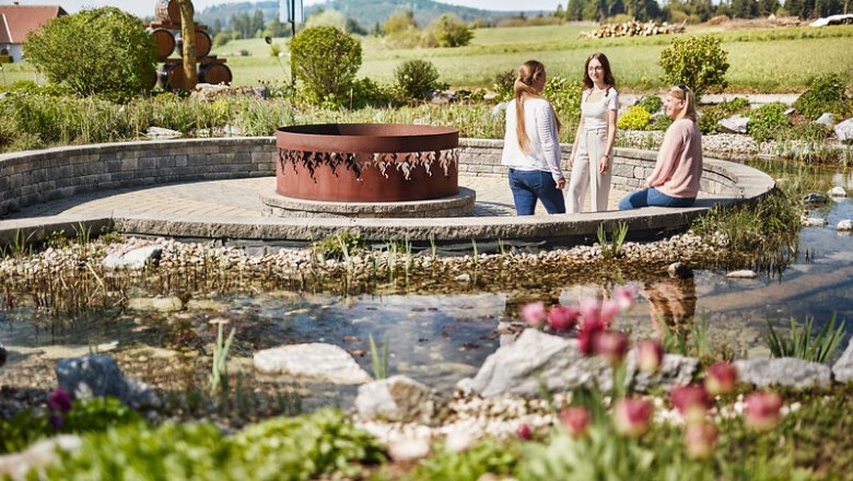 Drei Frauen sitzen in einem Garten mit einem kleinen Teich und einer Metallskulptur in der Mitte.