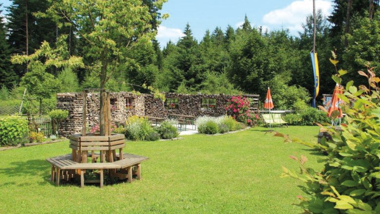 Ein grüner Garten mit Holzbänken um einen Baum, umgeben von Wald und Blumen.