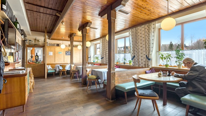 Gemütliche Gaststube mit Holzmöbeln und großen Fenstern, ein Mann sitzt an einem Tisch.