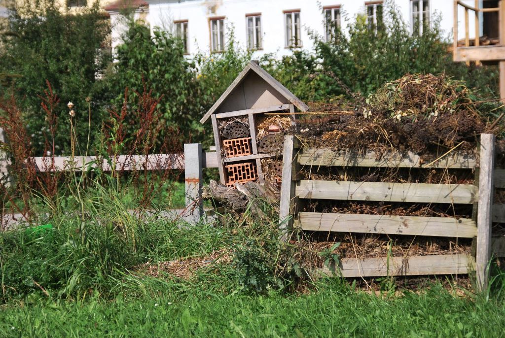 Ein Insektenhotel neben einem Komposthaufen in einem Garten mit grünem Gras und Bäumen im Hintergrund.