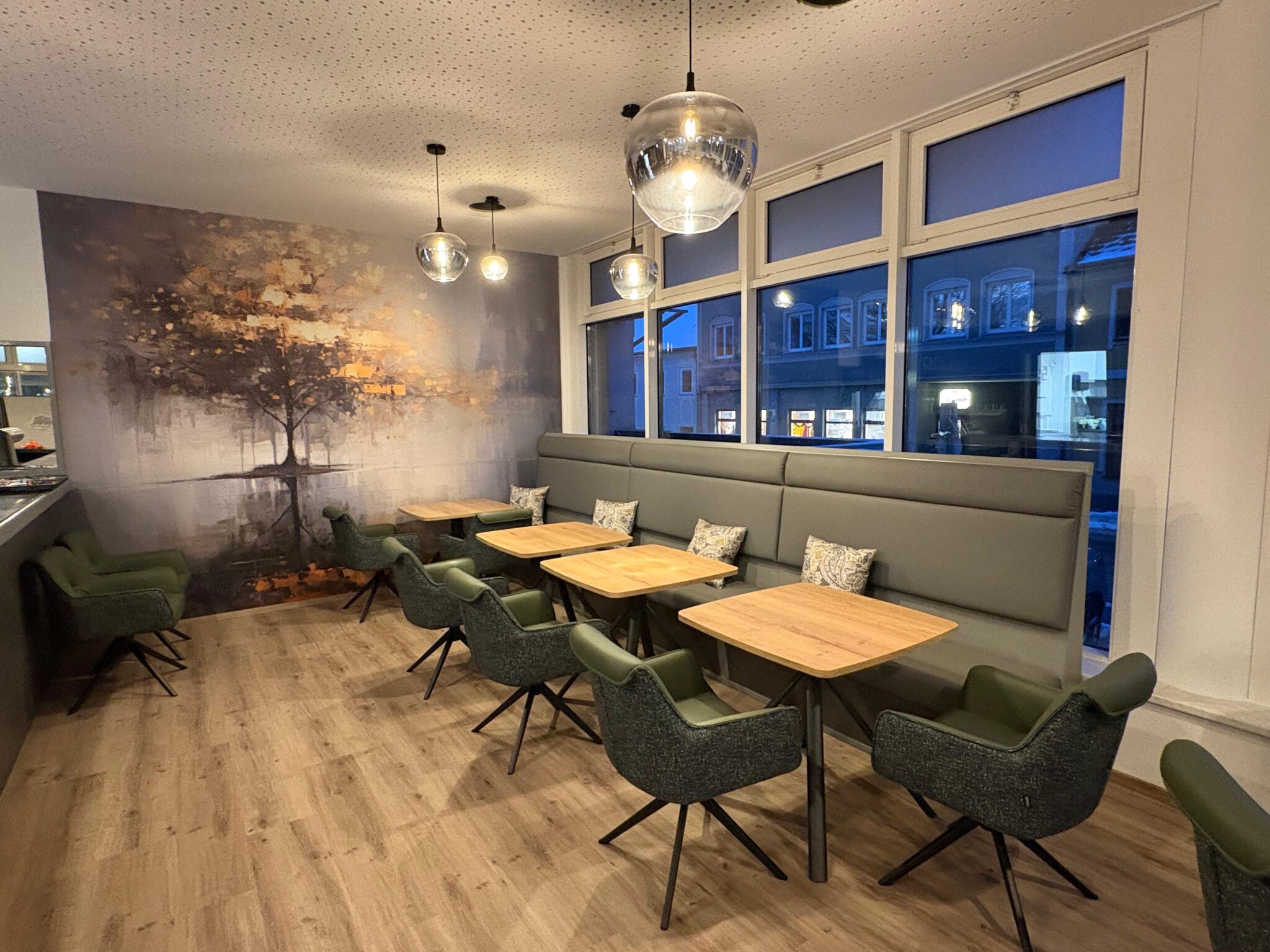 Gemütlicher Café-Bereich mit Sitzbänken, Tischen und Stühlen vor einer Wand mit Baumgemälde.