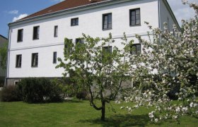 Ein weißes Gebäude mit braunem Dach und blühenden Bäumen im Vordergrund.