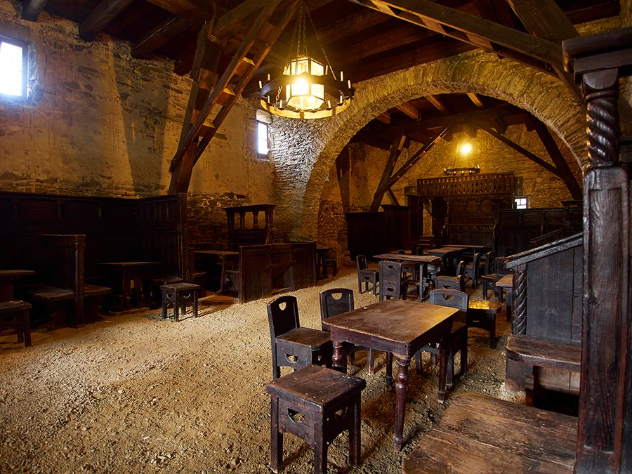 Innenansicht einer mittelalterlichen Taverne mit Holzmöbeln und Steinwänden.