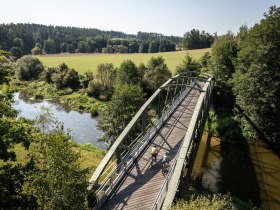 Die sanfte Br&uuml;cke schwingt &uuml;ber den glitzernden Fluss, umgeben von &uuml;ppigem Gr&uuml;n und der Ruhe der Natur. Radfahrer genie&szlig;en die frische Luft und die malerische Landschaft, w&auml;hrend die Sonne sanft auf die Wiesen scheint.