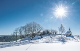 In dieser winterlichen Idylle strahlt die Sonne hell &uuml;ber die schneebedeckten H&uuml;gel und die charmante Kapelle. Die klare, kalte Luft und die glitzernde Schneedecke schaffen eine friedliche Atmosph&auml;re, die zum Verweilen einl&auml;dt. Fu&szlig;spuren im Schnee erz&auml;hlen von den Abenteuern der Besucher, die diese zauberhafte Landschaft erkunden.