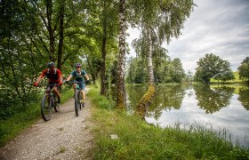 Zwei Mountainbiker genie&szlig;en die frische Luft und die malerische Landschaft entlang eines ruhigen Weges, der von hohen B&auml;umen ges&auml;umt ist. Der glitzernde See spiegelt die Wolken und das satte Gr&uuml;n der Umgebung wider, w&auml;hrend die beiden Radfahrer mit Freude und Energie in die Natur eintauchen.