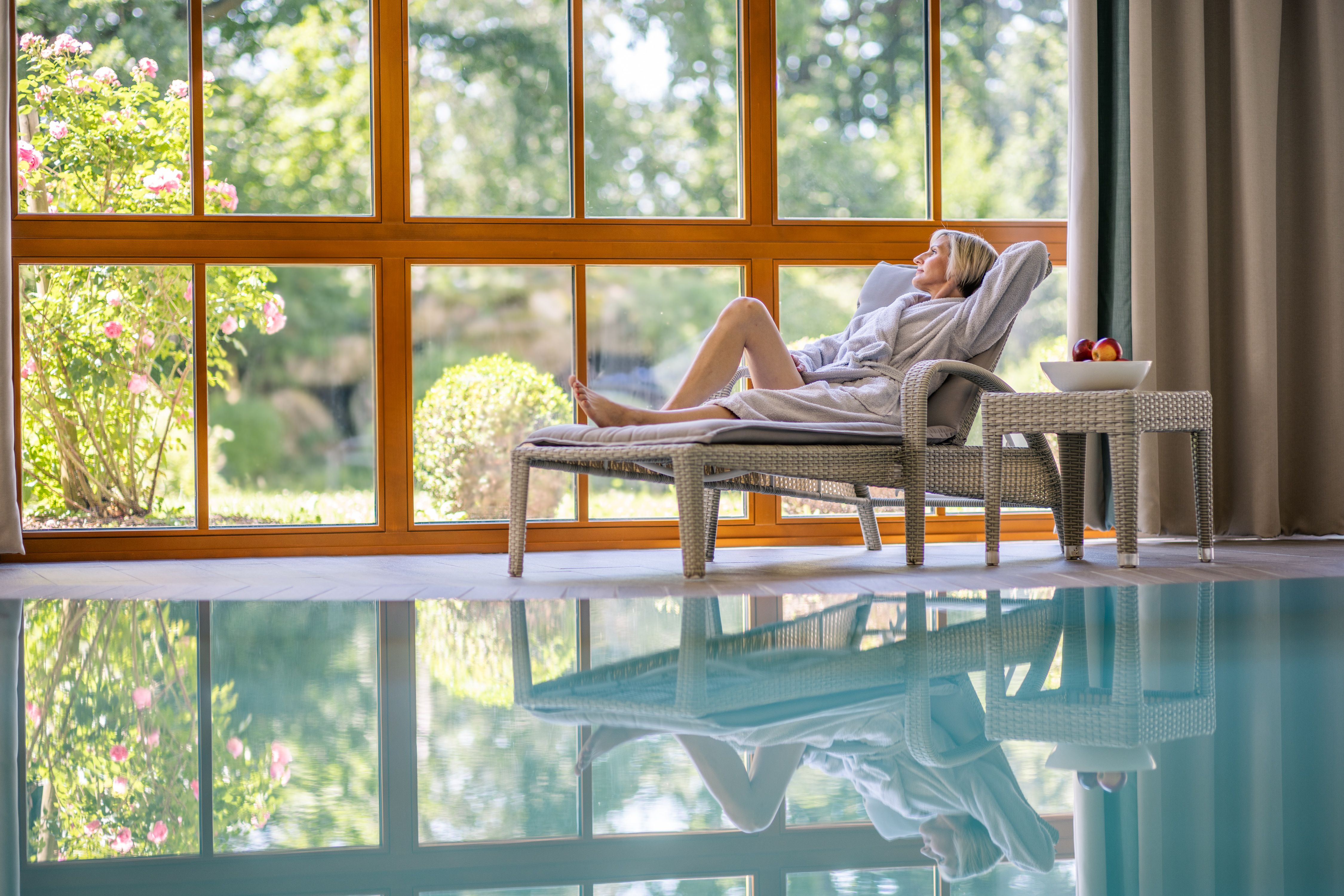 Eine Frau entspannt auf einer Liege in einem Spa mit Blick auf einen Garten durch große Fenster.