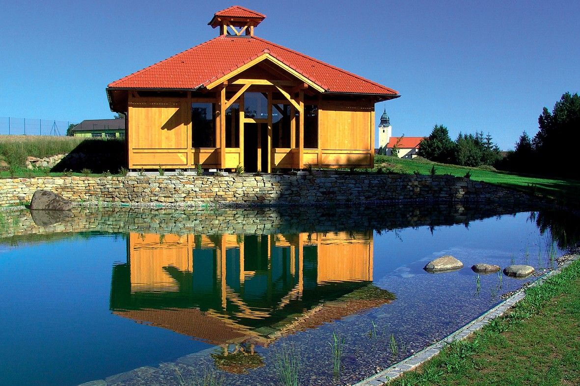 Holzgebäude mit rotem Dach spiegelt sich in einem Teich, umgeben von grüner Landschaft.