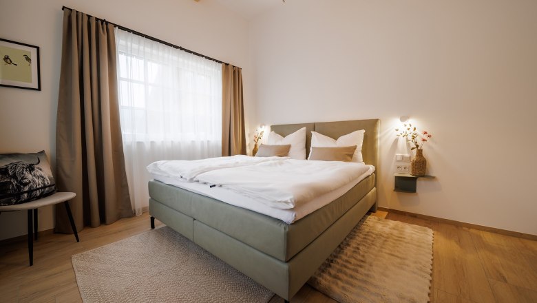 Modernes Schlafzimmer mit Doppelbett, Holzboden und großen Fenstern mit Vorhängen.