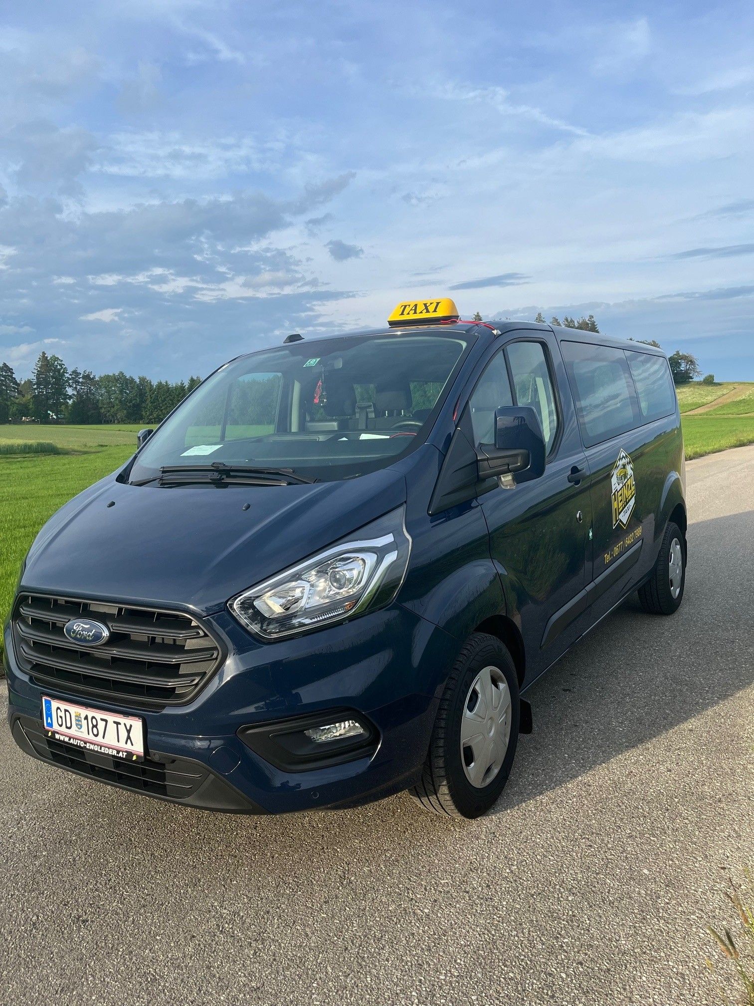 Ein blaues Ford-Taxi steht auf einer Landstraße vor einer grünen Wiese und einem bewölkten Himmel.