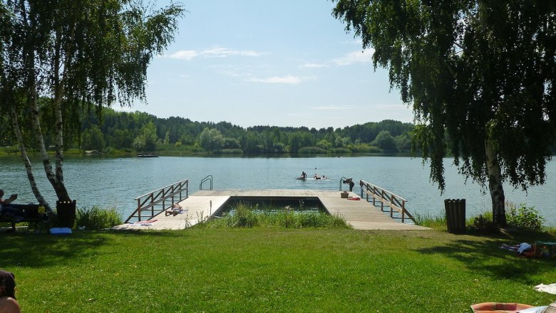 Ein Steg am Bergwerksee Langau mit Menschen, die schwimmen und sich sonnen. Umgeben von Bäumen und grüner Wiese.