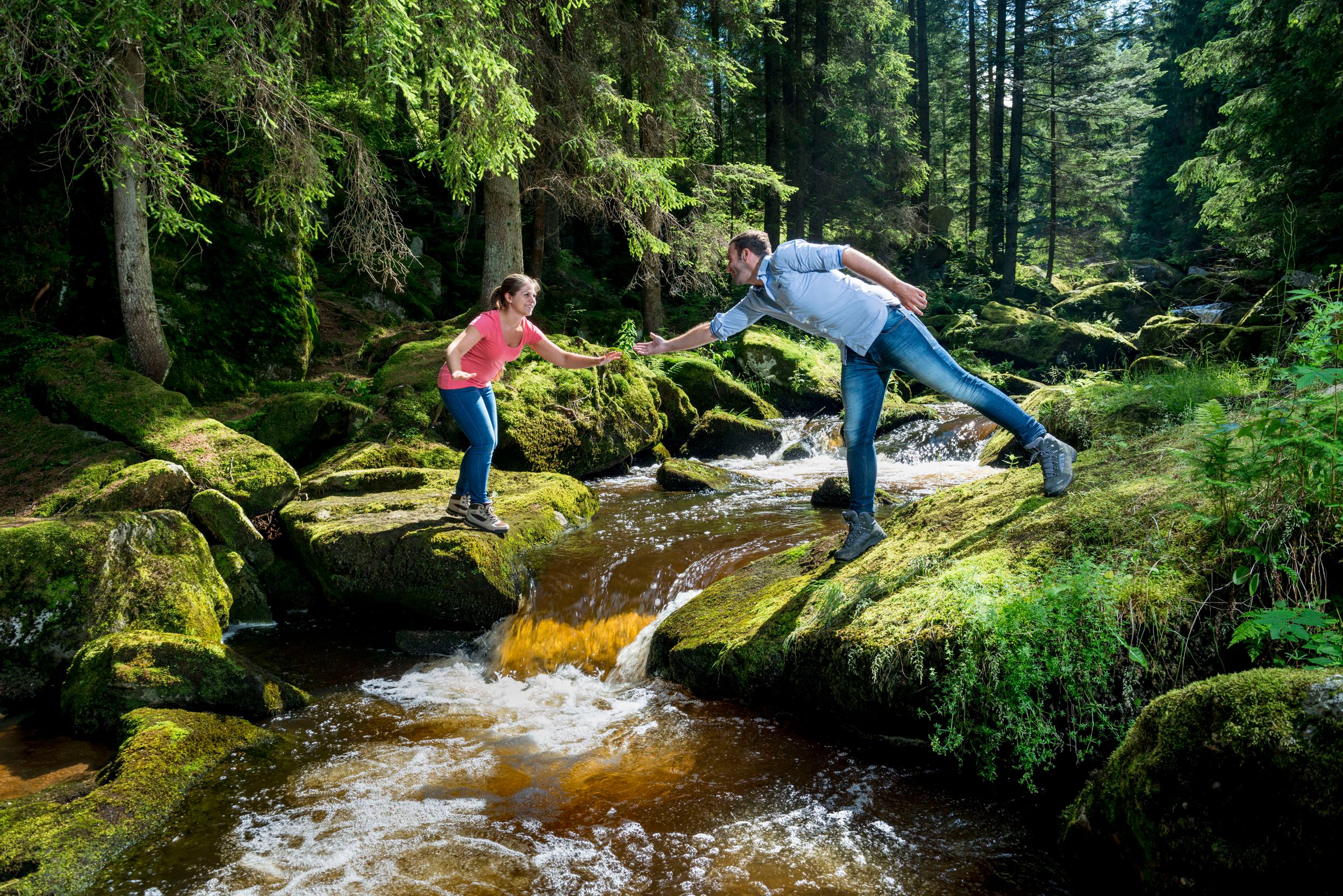 Zwei Personen stehen auf moosbedeckten Steinen über einem Bach in einem Wald und reichen sich die Hände.