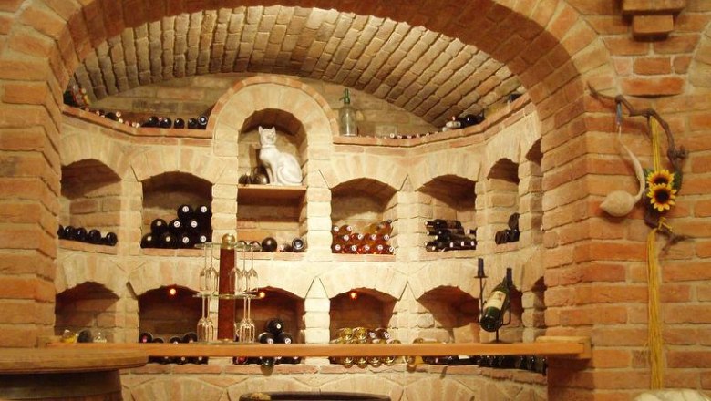 A wine cellar with brick shelves filled with bottles of wine. A cat figure stands in the middle. A sunflower hangs on the wall.