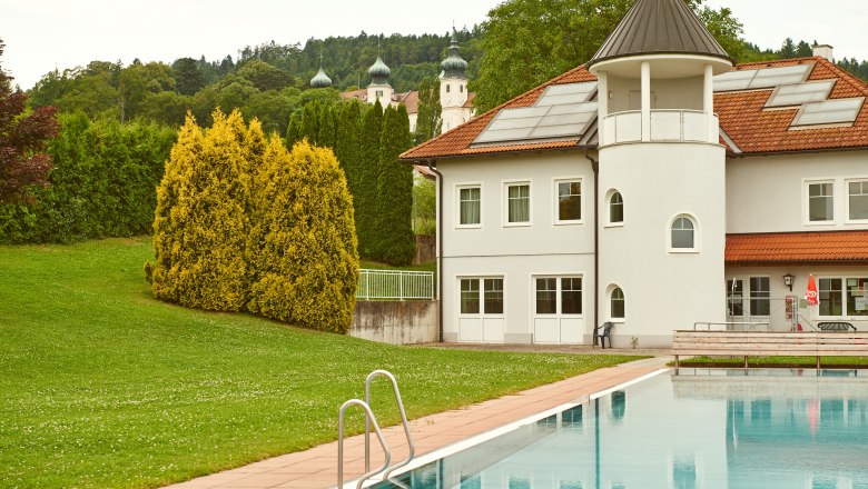 Außenansicht einer Freizeitanlage mit Pool und Gebäude, im Hintergrund Bäume und Kirchtürme.