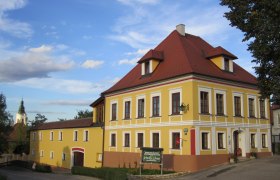 Gelbes zweistöckiges Gebäude mit rotem Dach und weißen Fensterrahmen, daneben ein kleineres Gebäude. Im Hintergrund eine Kirchturmspitze.