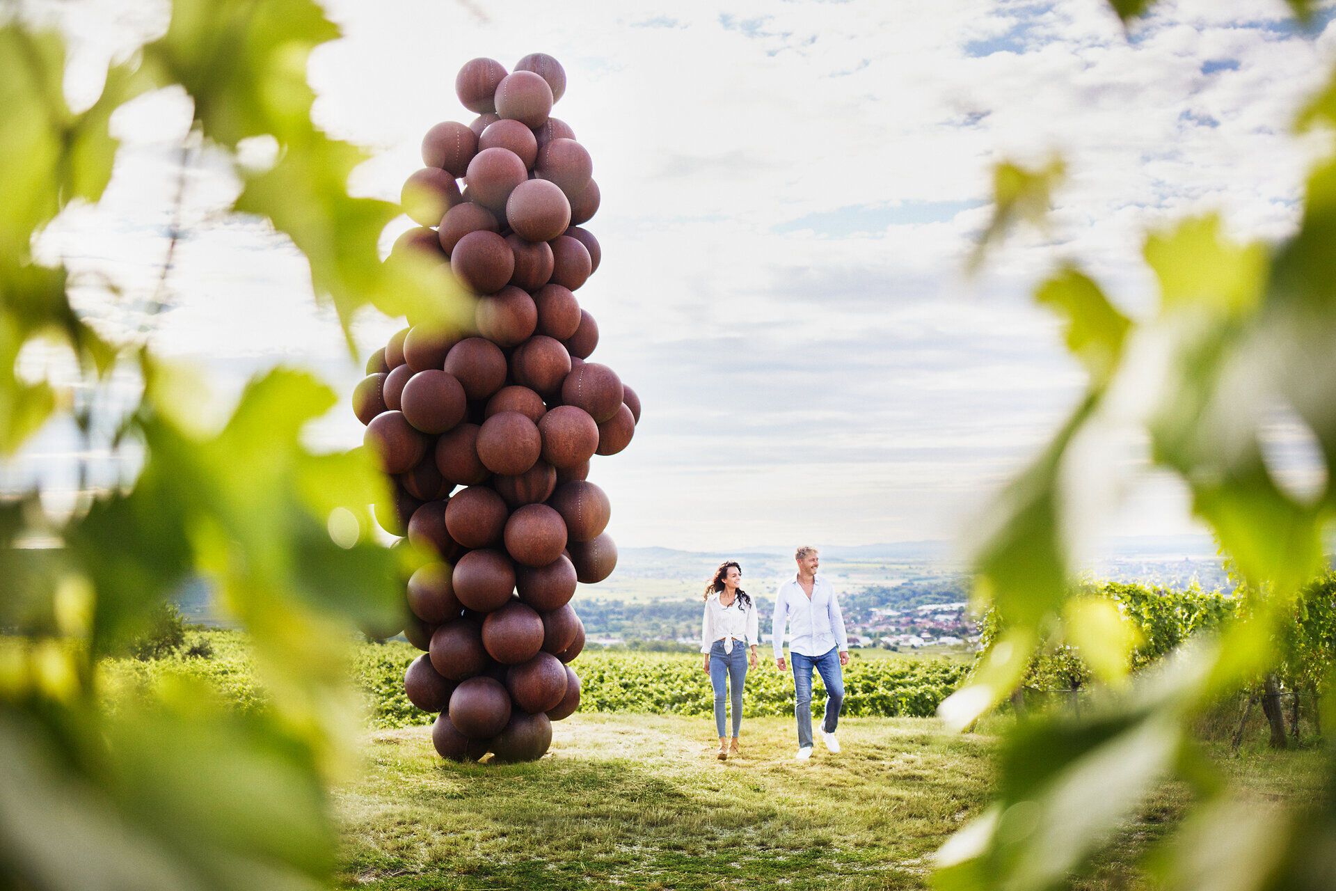 Inmitten der sanften Hügel des Kamptals schlendern zwei Personen entspannt durch die Weinberge, umgeben von üppigem Grün und der beeindruckenden Riesenweintraube, die als Kunstwerk die Landschaft ziert. Die frische Luft und die malerische Aussicht laden dazu ein, die Seele baumeln zu lassen und die Schönheit der Natur zu genießen.