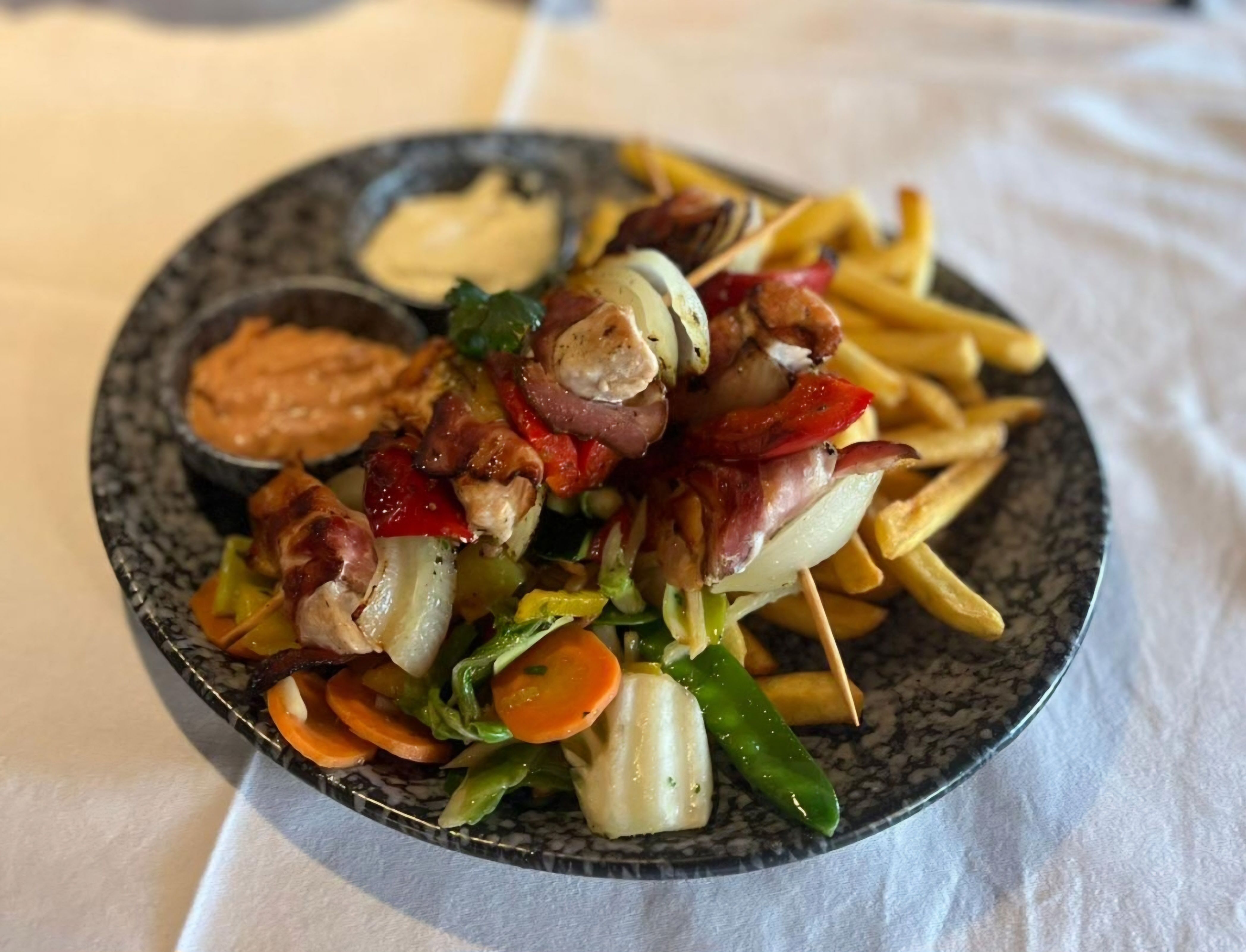 Ein Teller mit Pommes, Hühnerspießen mit Gemüse und zwei Dips auf einem Tisch.