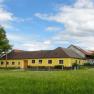 Gelbes Bauernhaus auf einer grünen Wiese mit blauem Himmel und Wolken.