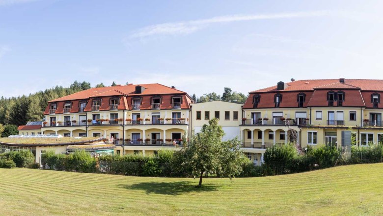 Außenansicht des Gesundheitshotels Moorbad Bad Großpertholz mit rotem Dach und Balkonen, umgeben von grüner Landschaft.