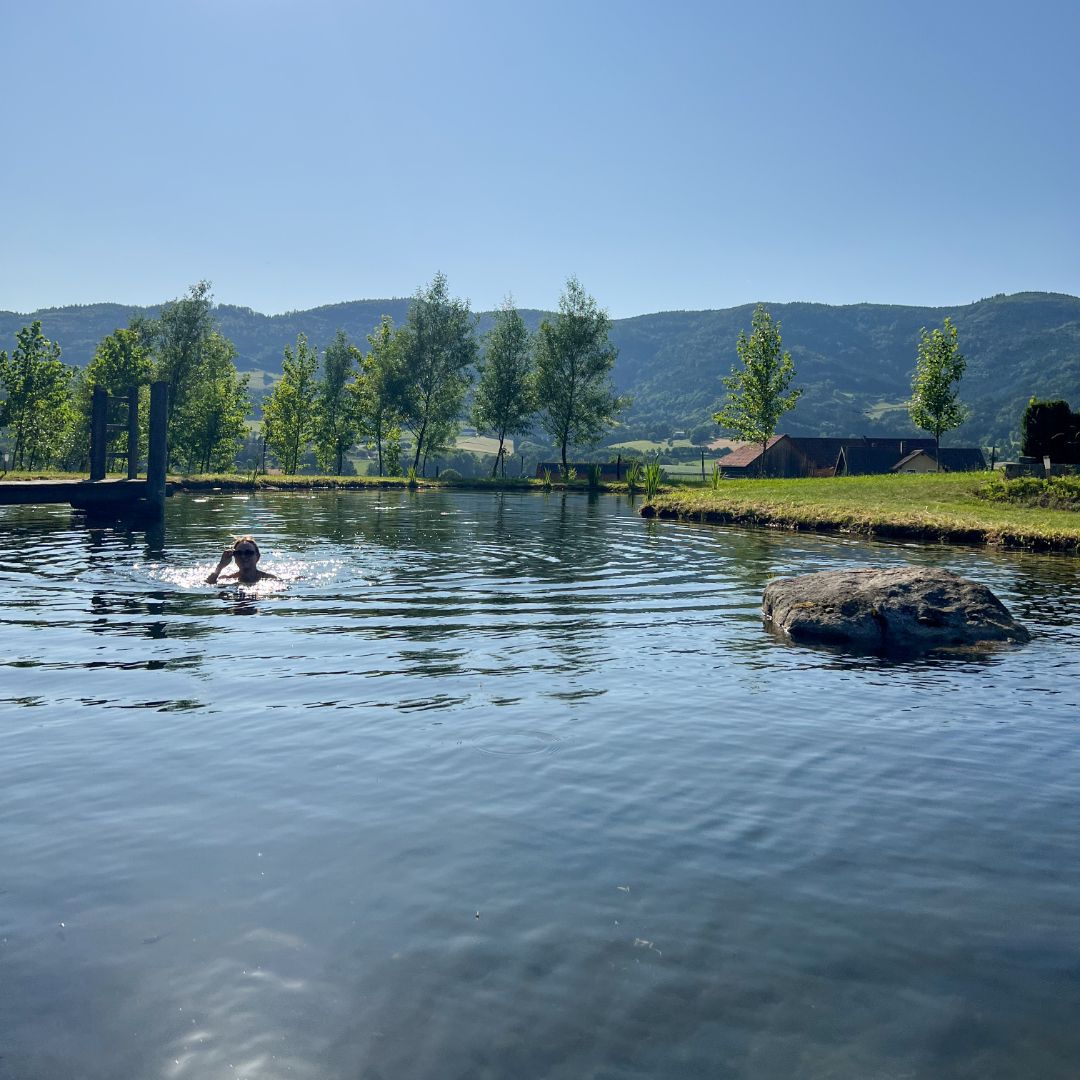 Person schwimmt in einem Teich mit Bäumen und Hügeln im Hintergrund.