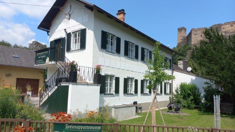 Apartments Zum Burgblick, © Jürgen Mokesch A white building with green shutters, surrounded by a garden, with a castle in the background.