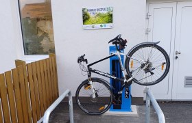 Fahrrad an einer blauen Service-Station befestigt, neben einem Geb&auml;ude mit einem Schild 'Fahrrad-Rastplatz & Service Station'.