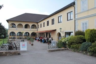 Ein gelbes Gebäude mit Arkaden und Terrasse, auf der Menschen sitzen.