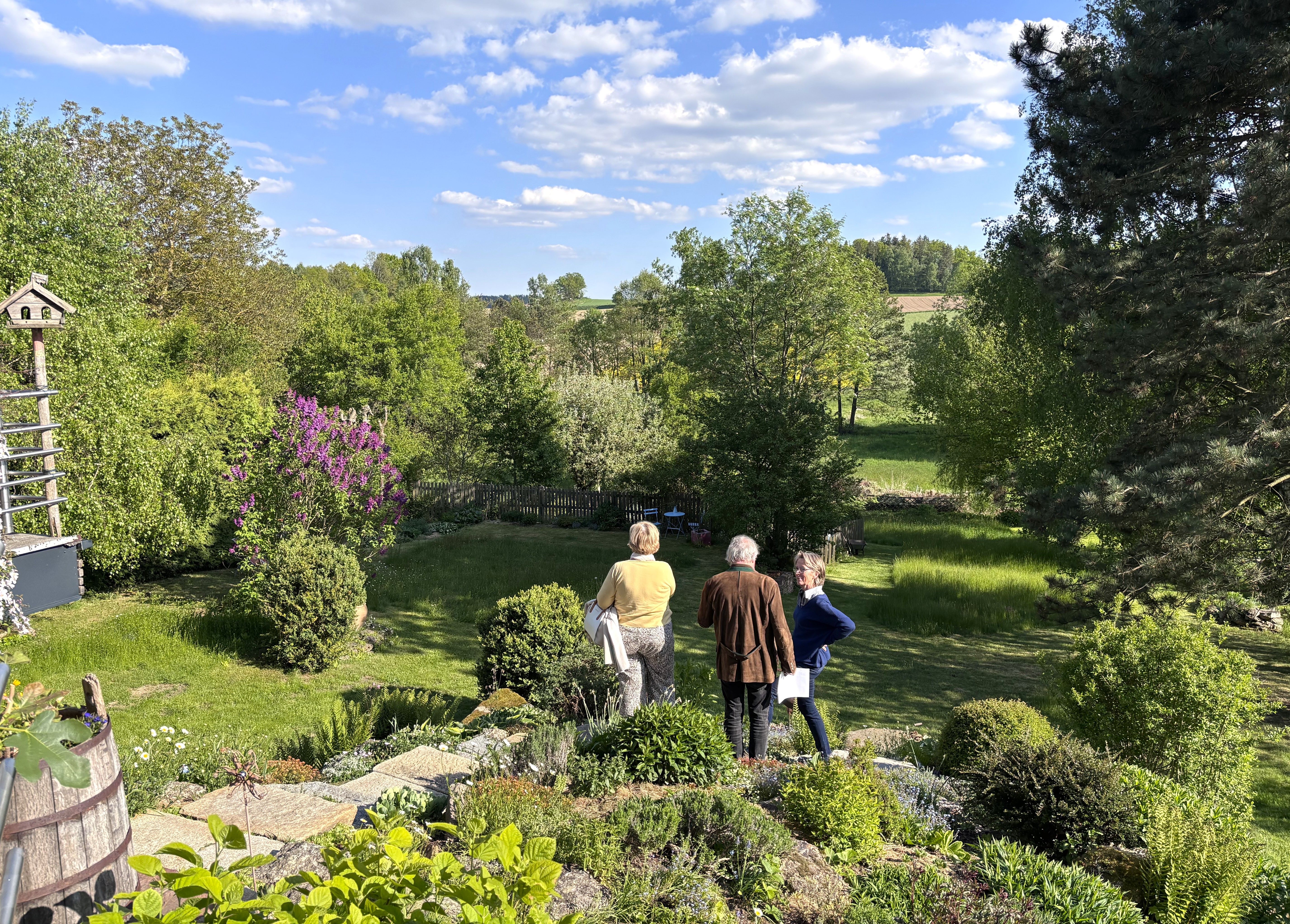 Drei Personen stehen in einem grünen Garten mit Bäumen und Sträuchern, umgeben von einer ländlichen Landschaft unter blauem Himmel mit Wolken.