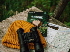 Einladend liegt die rustikale Bank im Grünen, umgeben von der frischen Luft der Natur. Die leuchtend gelbe Mütze und die hochwertigen Ferngläser laden dazu ein, die atemberaubenden Ausblicke zu genießen und die Schönheit der Umgebung zu erkunden. Eine Karte und ein Informationsheft versprechen spannende Abenteuer auf den Wanderwegen.