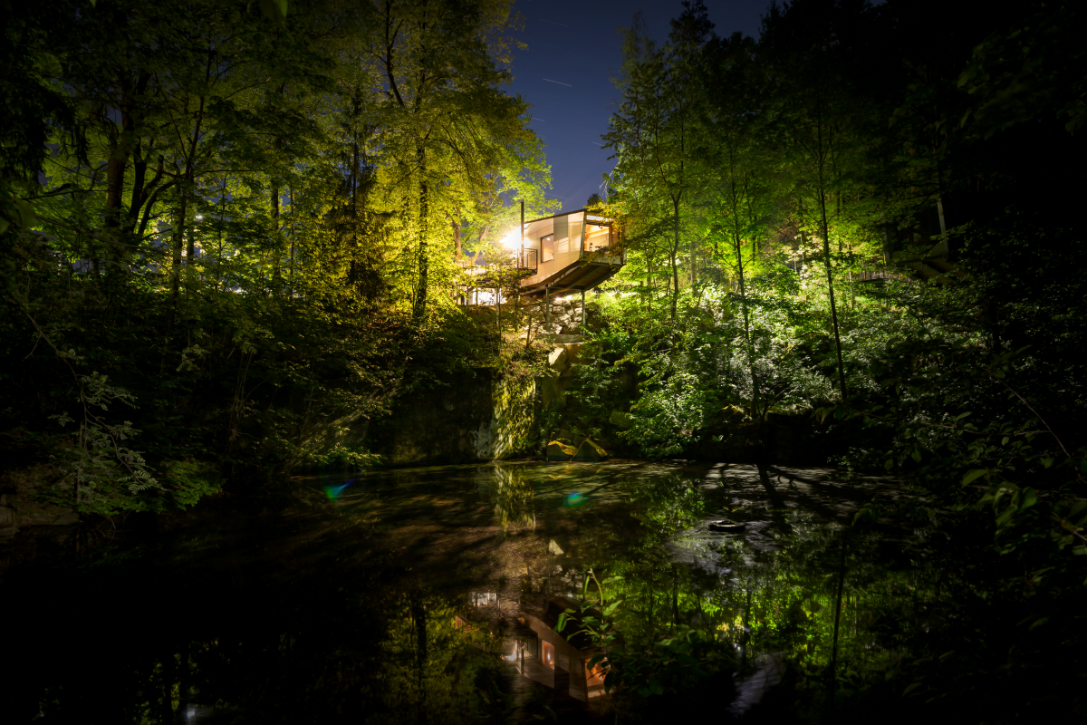 Beleuchtetes Baumhaus in einem Wald bei Nacht, reflektiert in einem Teich.