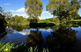 Ein ruhiger Fluss im Naturpark Dobersberg mit Bäumen und blauem Himmel im Hintergrund.