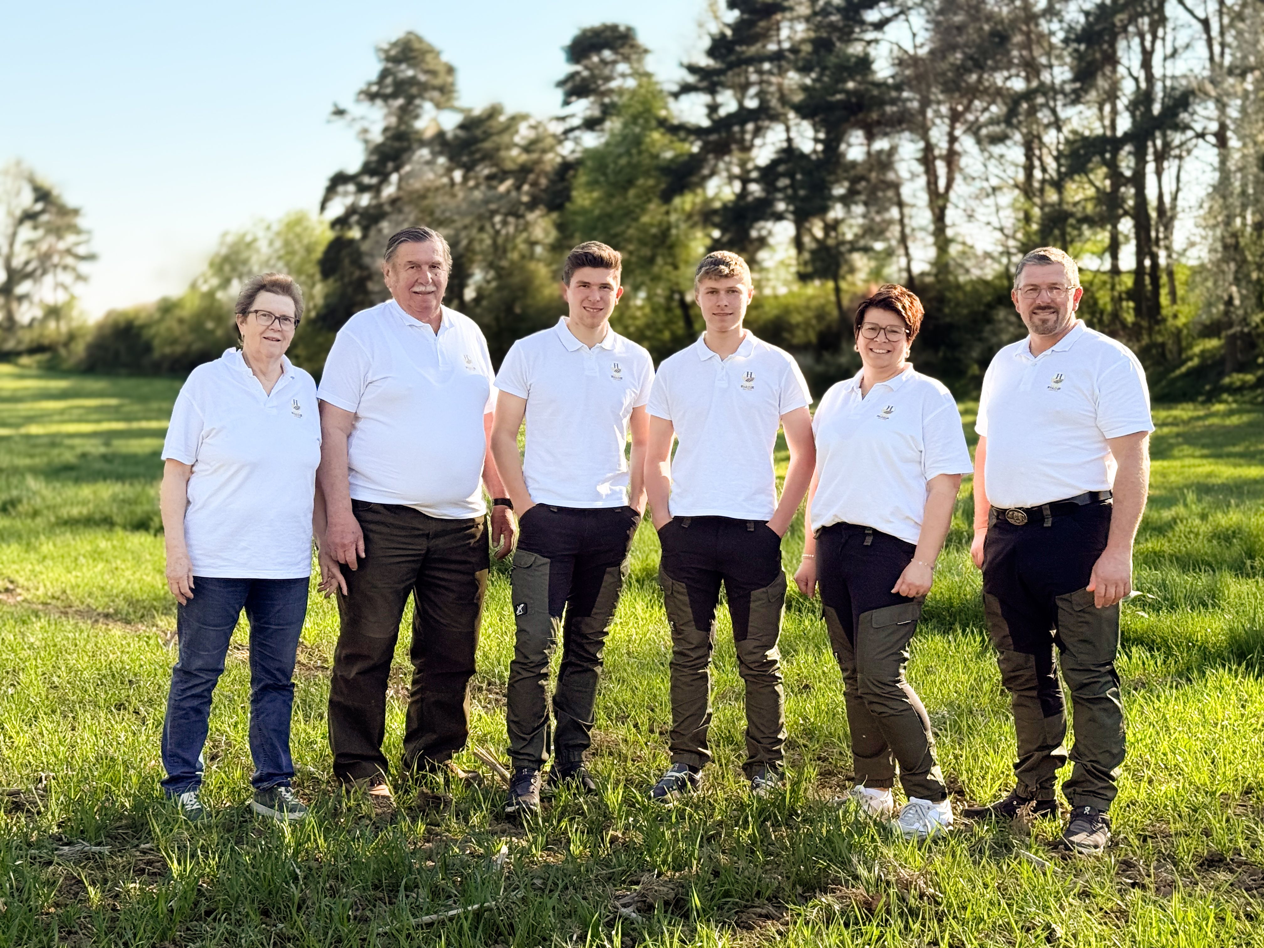 Sechs Personen in weißen Polohemden stehen auf einer Wiese vor Bäumen.