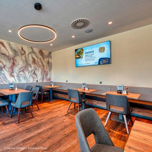 Modernes Restaurant mit Holzboden, grauen Stühlen und einem großen Bildschirm an der Wand.