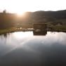Ein Teich mit einem Holzgebäude am Ufer, umgeben von Hügeln und Bäumen im Sonnenuntergangrgang.