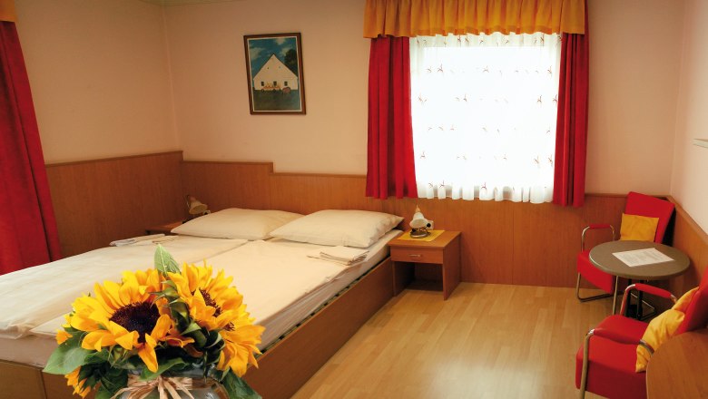 A cozy room with a double bed, red curtains, a table with chairs and a bouquet of sunflowers in the foreground.