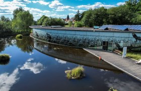 Geb&auml;ude mit bemalter Fassade und Solarpanelen, umgeben von Wasser und B&auml;umen.
